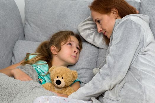 Happy mom sitting on couch with her preteen daughter. Smiling loving mother hugging her daughter lying with teddy bear toy. Woman and child wearing indoor clothes sitting together covered with plaid