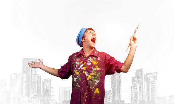 Young emotional artist with open mouth gesturing with paintbrush. Happy painter in shirt and bandana standing on background modern office buildings. Creative hobby and artistic occupation concept