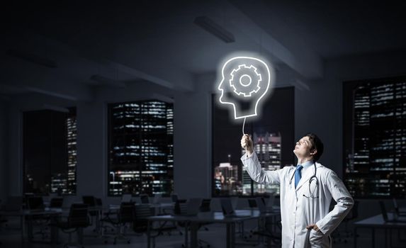 Conceptual image of confident doctor in white uniform interracting with glowing gear brain symbol while standing against night cityscape view on background.