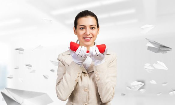 Smiling young woman holding retro red phone in office with flying paper planes. Call center operator in business suit with landline phone. Hotline telemarketing. Business assistance and consultation.