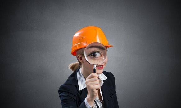 Technical inspector looking through magnifying glass. Woman civil engineer in orange safety helmet checking with magnifier on grey wall background. Commercial real estate appraisal and consultation.