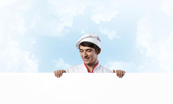 Young male chef with blank signboard. Handsome chef in white and red uniform on blue sky background. Cooking classes announcement. Professional catering service concept with copy space.