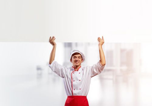 Young male chef with blank signboard above head. Chef in white and red uniform on light room blurred background. Cooking classes announcement. Professional catering service concept with copy space