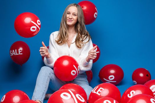 caucasian woman with attractive appearance sits with baloons with percent print and smiles, picture isolated on blue background