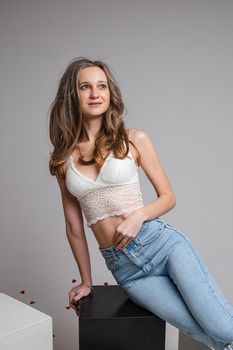 Pretty slim long haired caucasian girl in white top lingerie, blue jeans posing for studio portrait on gray background. High quality photo