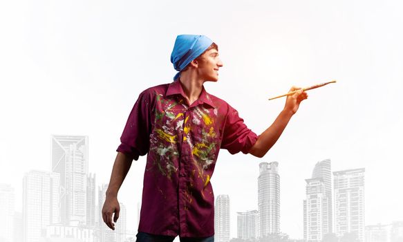 Young male artist holding paintbrush. Happy painter in dirty shirt and bandana standing on background modern cityscape. Creative hobby and artistic occupation concept with copy space