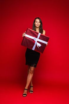 Full length portrait of attractive Caucasian woman in black skirt and sandals holding big wrapped gift with white bow. Isolated on red.