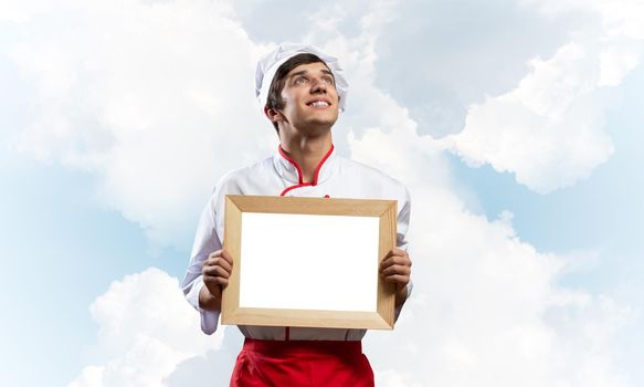 Young male chef holding blank white board for text. Handsome chef in white and red uniform on blue sky background. Cooking classes advertising. Professional catering service concept with copy space.