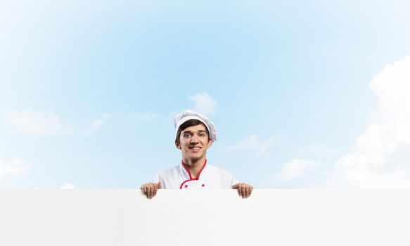 Young male chef with blank signboard. Handsome chef in white and red uniform on blue sky background. Cooking classes announcement. Professional catering service concept with copy space.