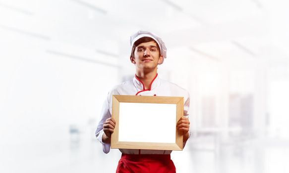 Young male chef standing near cooking table and holding blank white board. Handsome chef in white and red uniform in light kitchen interior. Cooking classes advertising. Professional catering service