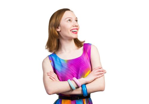 Smiling woman standing with folded arms and looking up aside. Elegant fashionable lady wears bright dress and bracelets. Studio shot of joyful redhead girl with perfect teeth and healthy clean skin.