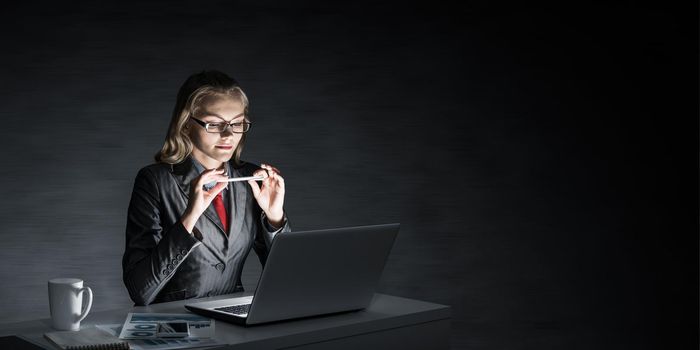 Attractive young businesswoman in glasses sitting at office desk and working at laptop. Beautiful lady in business suit and tie busy at work in evening. Career building and business development