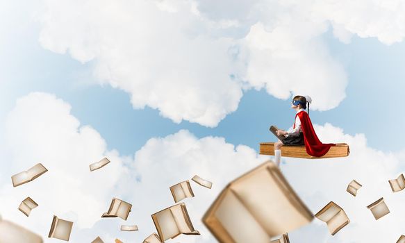 Little child in mask and cape floating on book in sky and reading