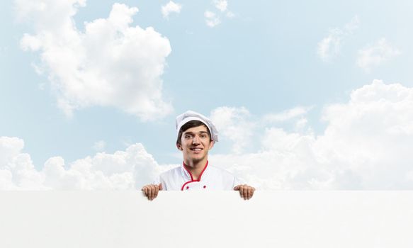 Young male chef with blank signboard. Handsome chef in white and red uniform on blue sky background. Cooking classes announcement. Professional catering service concept with copy space.