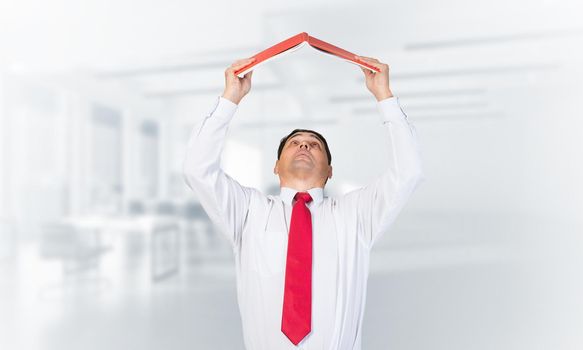Senior businessman holding open book overhead as roof. Portrait of adult man in white shirt and tie standing in blurred office interior. Education and professional knowledges. Reference information.