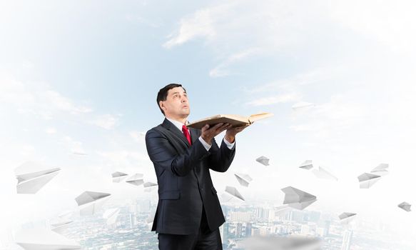 Serious businessman with open book looking up. Man in suit standing on downtown background with flying paper planes. Lawyer holding big legal regulation book. Business accounting and consulting.