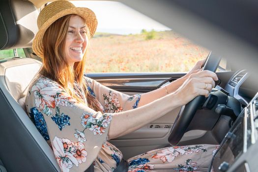 A smiling woman in a hat and a colored dress is sitting behind the wheel of a car, strapped in, outside the window is a large blooming field of poppies.