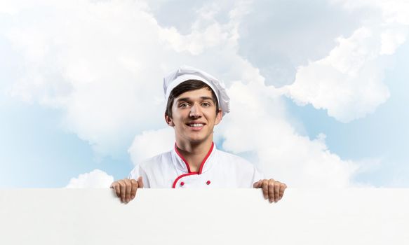 Young male chef with blank signboard. Handsome chef in white and red uniform on blue sky background. Cooking classes announcement. Professional catering service concept with copy space.