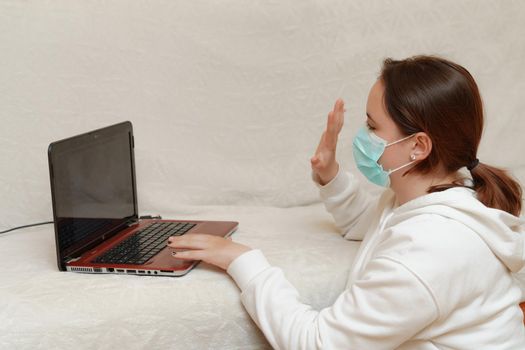 A young girl in a mask lies at home on a sofa with a computer and communicates via skype with friends, makes gestures with her hands. covid-19 ehidemic concept. Quarantine Stay at home