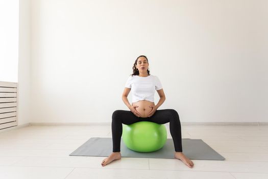 A young pregnant woman doing relaxation exercise using a fitness ball while sitting on a mat and holding her tummy