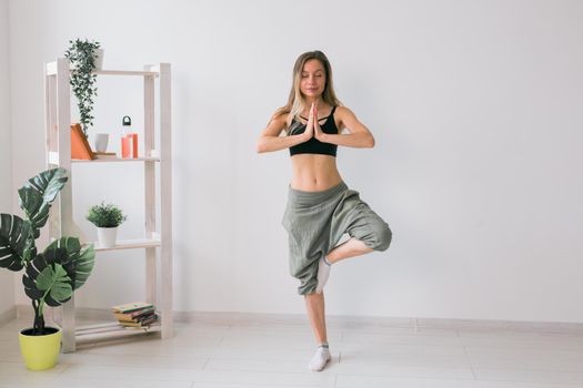 Woman practise yoga performs. Tree pose standing on mat inside of cozy room with plants and greenery. Healthy lifestyle