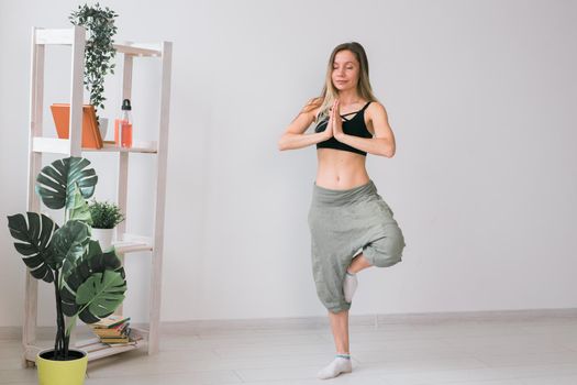 Woman practise yoga performs. Tree pose standing on mat inside of cozy room with plants and greenery. Healthy lifestyle