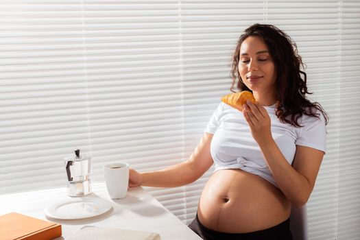 Joyful pregnant young beautiful woman eating croissant and drinking tea during morning breakfast. Concept of pleasant morning and expectation of meeting baby.