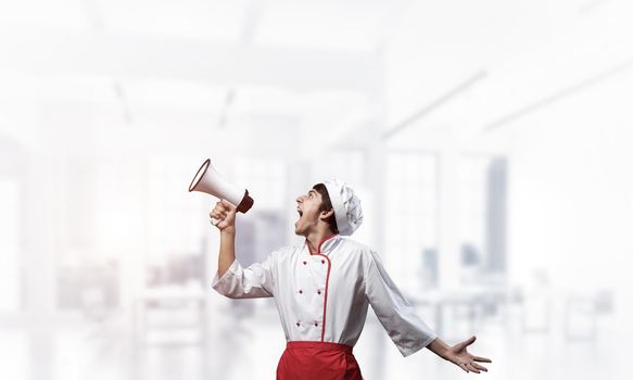 Young chef shouting loudly into megaphone. Emotional caucasian chef in white hat and red apron on light blurred background. Restaurant advertisement, food retail campaign announcement.