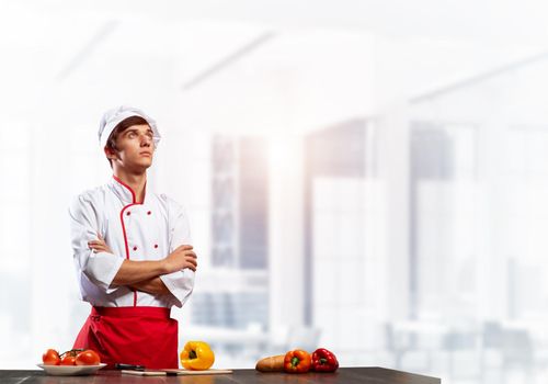 Young male chef standing with folded arms near cooking table with vegetables. Handsome chef in white hat and red apron on light room blurred background. Professional cooking classes advertising.