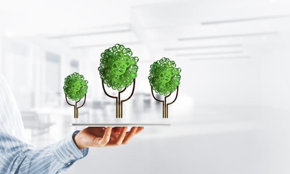 Close of businessman holding tablet pc with green tree made of gears and cogwheels. Mixed media