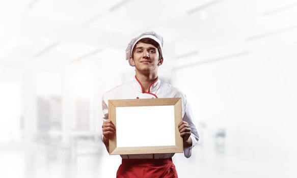 Young male chef standing near cooking table and holding blank white board. Handsome chef in white and red uniform in light kitchen interior. Cooking classes advertising. Professional catering service