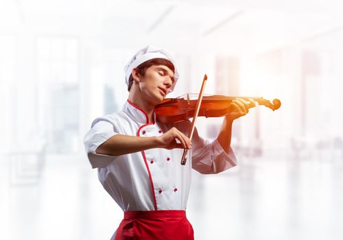 Young male chef with violin standing near cooking table with vegetables. Handsome chef in white hat and red apron in light kitchen interior. Creative culinary performance. High cooking skills concept