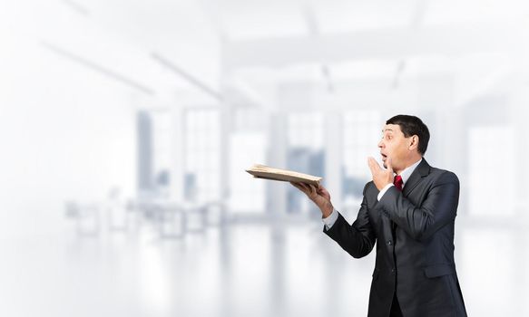 Scared businessman covering mouth with hand and holding open book. Startled adult man in business suit and tie standing on blurred office background. Education and knowledges. Business accounting