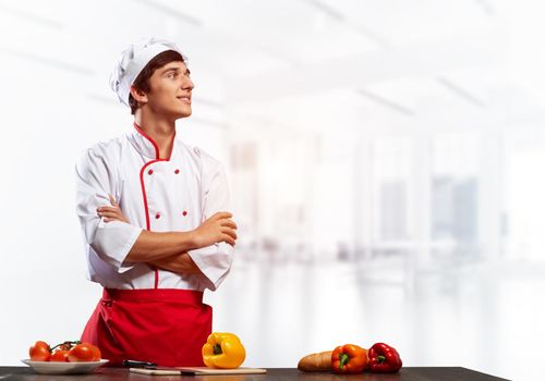 Young male chef standing with folded arms near cooking table with vegetables. Handsome chef in white hat and red apron on light room blurred background. Professional cooking classes advertising.
