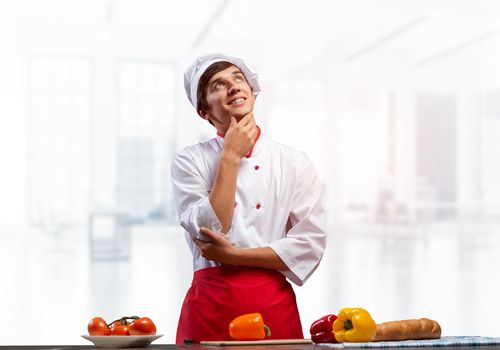 Young male chef standing with folded arms and looking up dreamily. Pensive culinary school student in white hat and red apron in light kitchen interior. Professional kitchen staff recruitment