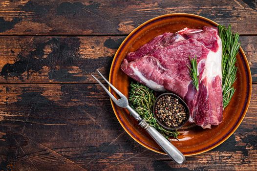 Raw lamb mutton thigh or leg with rosemary and thyme in rustic plate. Dark wooden background. Top view. Copy space.