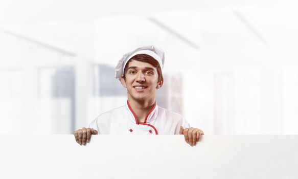 Young male chef with blank white signboard. Handsome chef in white and red uniform on light room blurred background. Cooking classes announcement. Professional catering service concept with copy space