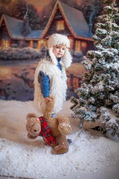 A boy in a white hat and boots on the background of an artificial forest in the winter village