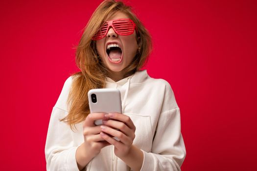 Beautiful emotional young blonde woman wearing stylish white hoodie and funny colorful glasses standing isolated over colourful background holding and using mobile phone communicating online on the internet and yawning.
