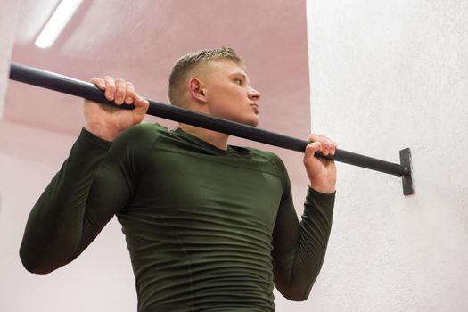 Young sports guy in a green thermal suit pulls himself up on the horizontal bar in the gym.