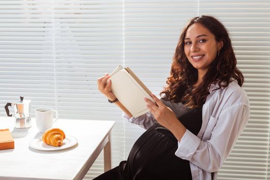 Pregnant woman eating and reading book at home. Morning, breakfast and pregnancy concept.