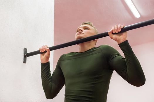 Young sports guy in a green thermal suit pulls himself up on the horizontal bar in the gym.