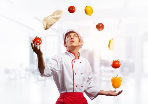 Young male chef juggles with food ingredients. Handsome chef in white hat and red apron in light kitchen interior. Professional cooking classes advertising. Food retail campaign announcement.