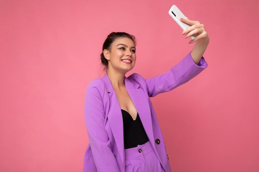 Photo of pretty young woman wearing purple suit taking selfie photo on the mobile phone isolated over pink background looking at phone camera.Copy space