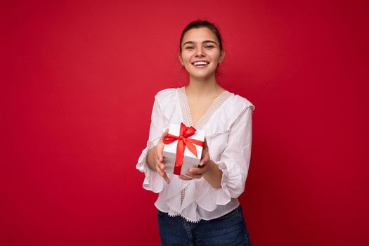 Beautiful happy young brunette woman isolated over colourful background wall wearing stylish casual clothes holding gift box and looking at camera. Empty space