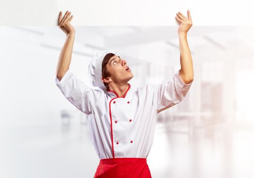 Young male chef with blank signboard above head. Chef in white and red uniform on light room blurred background. Cooking classes announcement. Professional catering service concept with copy space