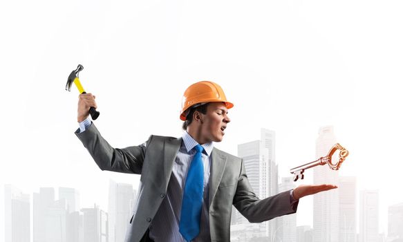 Furious businessman going to crash with hammer steel key. Young handsome contractor in business suit and safety helmet standing on cityscape background. Financial crisis and bankruptcy in real estate