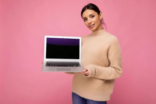 beautiful smiling young woman with dark hair looking at camera holding computer laptop with empty monitor screen with mock up and copy space wearing light colour sweater isolated on pink wall background.