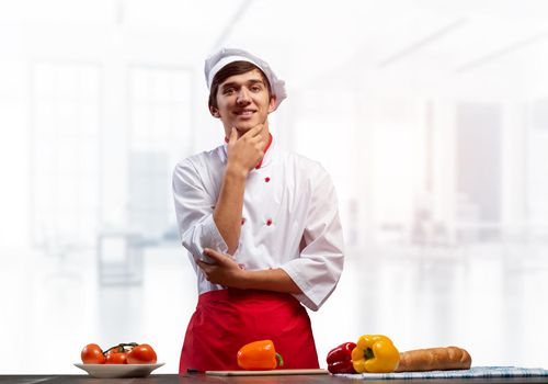 Young smiling chef standing near cooking table with fresh vegetables. Pleased chef in white hat and red apron in light kitchen interior. Cooking classes advertising. Restaurant food preparation