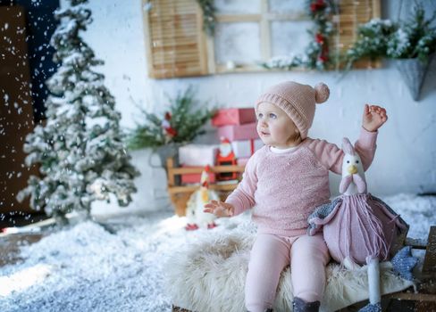 A baby girl sits in a Studio with artificial winter scenery in a pink sweater and a pink beanie.